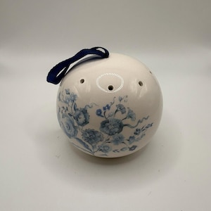 May include: A spherical white ceramic ornament featuring a blue floral pattern painted on the surface. The object has a series of small circular holes near the top and includes a dark blue fabric ribbon loop attached for hanging purposes.