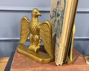 May include: A gold-toned metal eagle bookend with detailed feather textures, standing on a rectangular base. The eagle is positioned next to a vintage book with a blue and white cover illustration, resting on a stack of two hardcover books.