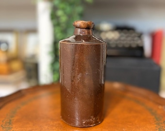May include: A cylindrical brown stoneware bottle with a glossy finish and a narrow neck. The base of the bottle features an embossed stamp that reads Doulton Lambeth. The bottle has a simple, straight-sided design with a slightly flared rim.