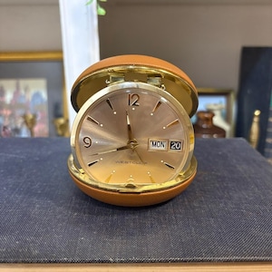 May include: A round Westclox travel alarm clock with a tan leather-like case that opens to reveal a gold-toned metallic face. The clock features gold hands, gold hour markers, and a rectangular window displaying the day MON and the date 20.