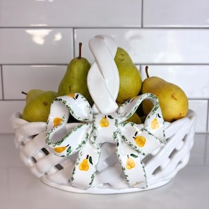 Peut inclure: Un panier en céramique blanche au design tressé avec une anse centrale torsadée. Un ruban décoratif en céramique, orné d'une bordure de vigne verte et d'illustrations de poires jaunes, est fixé à l'avant. Le panier contient plusieurs poires vertes.