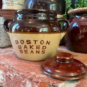 May include: A two-handled ceramic bean pot with a glossy dark brown glazed rim and lid, and a matte beige lower body. The text BOSTON BAKED BEANS is printed in dark brown capital letters on the front of the pot, which has a rounded shape.