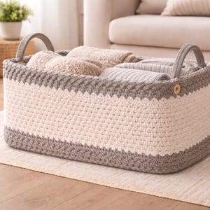 Puede incluir: Una cesta de almacenamiento rectangular, tejida a mano, en crema y gris. La cesta tiene dos asas arqueadas y está llena de mantas y textiles doblados. El diseño presenta un patrón de puntada texturizada y un detalle de botón.