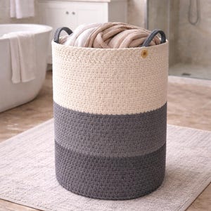 Peut inclure: Un panier à linge cylindrique tissé en trois tons, placé dans une salle de bain. Le panier est principalement crème en haut, gris au milieu et gris foncé en bas. Il est rempli de textiles pliés et possède des poignées grises.