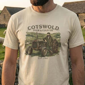 Op de afbeelding: Beige t-shirt met een afbeelding van een vintage tractor en een man in een landelijke omgeving. De tekst "Cotswold Farm & Country" en "EST. 1966" staan respectievelijk boven en onder de afbeelding. Het ontwerp roept een landelijk thema op.