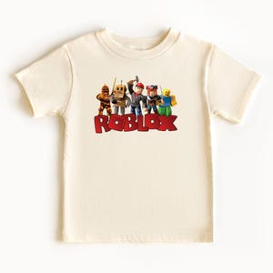 May include: Cream-colored t-shirt featuring the word "ROBLOX" in red block letters. The shirt displays a group of cartoon characters above the text. The characters are in various colors and poses, suggesting a playful theme.