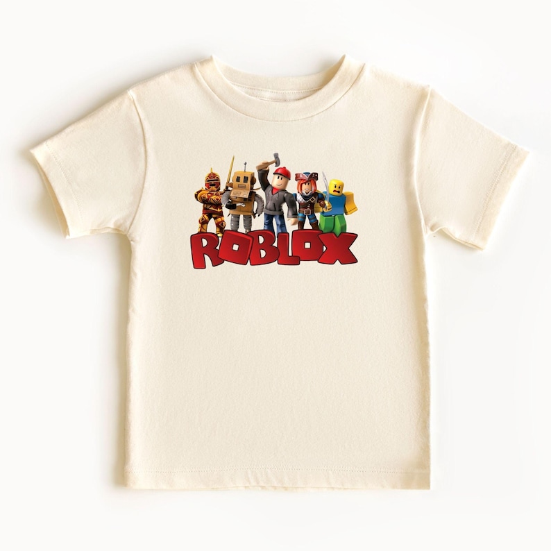 May include: Cream-colored t-shirt featuring the word "ROBLOX" in red block letters. The shirt displays a group of cartoon characters above the text. The characters are in various colors and poses, suggesting a playful theme.