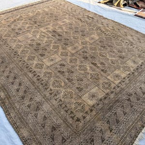 May include: A large, rectangular, vintage rug with a detailed geometric pattern in shades of brown and beige. The rug features a central design with repeating motifs and a decorative border. Fringes are visible along the edges.