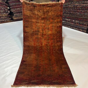 May include: A rectangular, vintage rug with a distressed appearance. The rug features a rich colour palette of browns, reds, and oranges, with intricate patterns and a fringed edge. The rug is held up by a person.