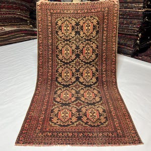 May include: A rectangular, patterned rug with a central design of repeating geometric shapes in shades of brown, orange, and beige against a black background. The border is a reddish-brown with intricate detailing. The rug is displayed on a white surface.