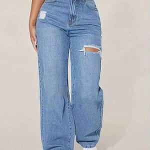 May include: Light blue denim jeans with a high waist and a relaxed, wide-leg fit. The jeans feature distressed details with rips at the knees and thighs. The model is wearing white sneakers.