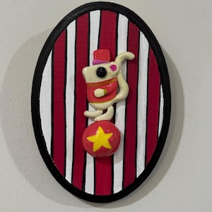 Puede incluir: Decoraci&oacute;n mural ovalada que representa un rat&oacute;n de circo haciendo equilibrio sobre una bola roja con una estrella amarilla. El fondo tiene rayas rojas y blancas, enmarcadas por un borde negro. El rat&oacute;n lleva un sombrero y una chaqueta rojos.