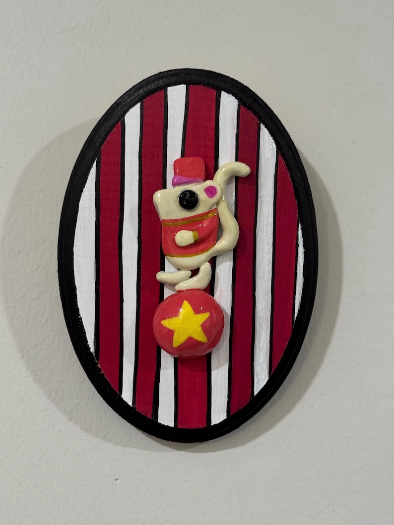 Puede incluir: Decoraci&oacute;n mural ovalada que representa un rat&oacute;n de circo haciendo equilibrio sobre una bola roja con una estrella amarilla. El fondo tiene rayas rojas y blancas, enmarcadas por un borde negro. El rat&oacute;n lleva un sombrero y una chaqueta rojos.