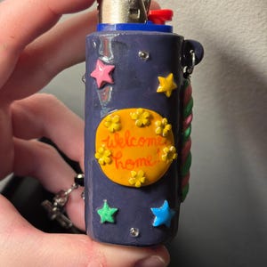 May include: A navy blue lighter decorated with colorful stars and flowers. The lighter has the words "Welcome Home!" on an orange oval. A decorative rope design is on the side, and a small chain is attached.