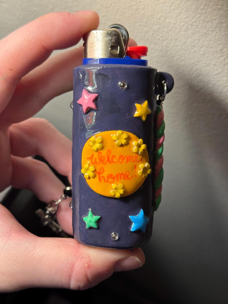 May include: A navy blue lighter decorated with colorful stars and flowers. The lighter has the words "Welcome Home!" on an orange oval. A decorative rope design is on the side, and a small chain is attached.