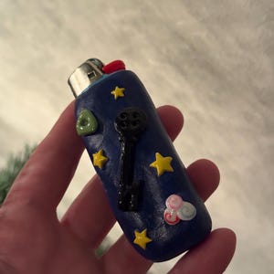 May include: A blue lighter decorated with yellow stars, a black key, a green triangle, and a pink and white cloud. The lighter has a red button and a silver metal top.