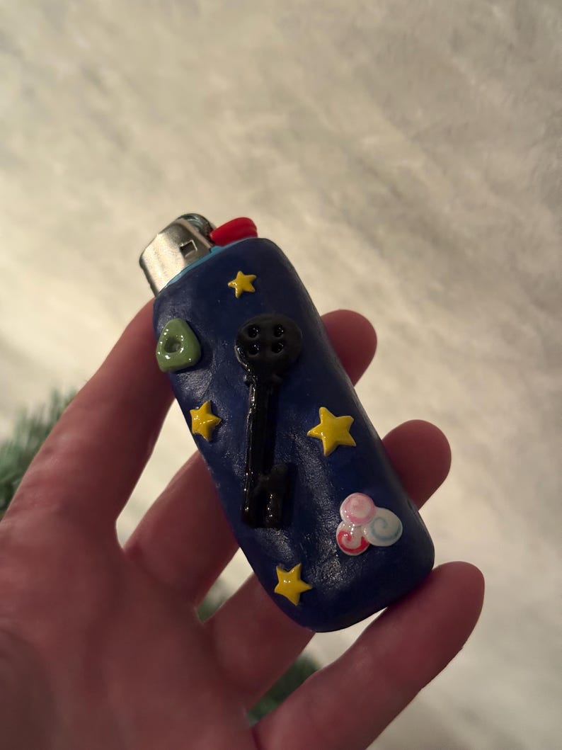 May include: A blue lighter decorated with yellow stars, a black key, a green triangle, and a pink and white cloud. The lighter has a red button and a silver metal top.