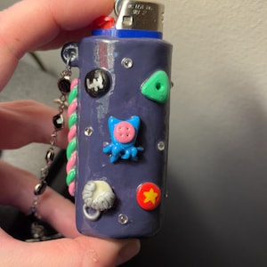 May include: A purple lighter decorated with various colorful embellishments, including a green triangle, a red star, and a blue octopus. The lighter has a silver chain with black beads and a pink and green twisted cord.