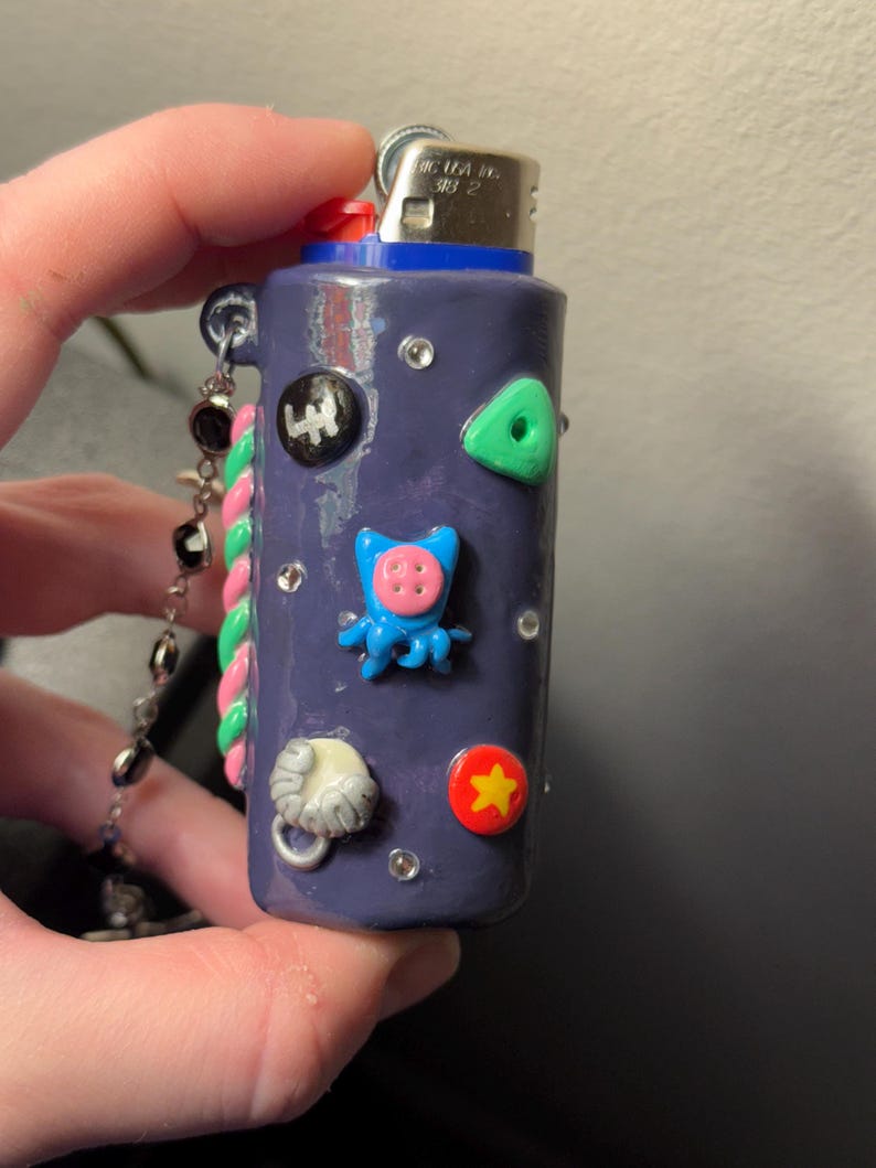 May include: A purple lighter decorated with various colorful embellishments, including a green triangle, a red star, and a blue octopus. The lighter has a silver chain with black beads and a pink and green twisted cord.