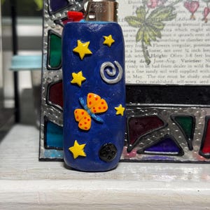 May include: A blue lighter decorated with yellow stars, a butterfly, and a swirl design. The lighter has a red ignition button and a silver metal top. The design is hand-crafted and whimsical.