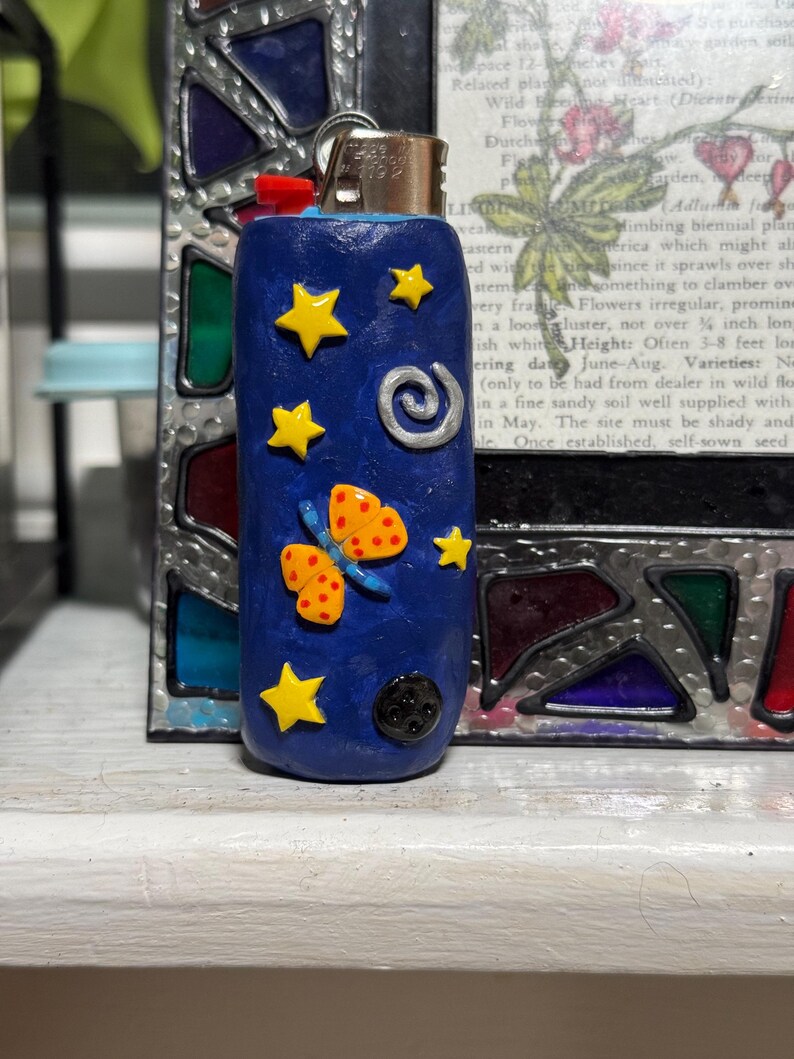 May include: A blue lighter decorated with yellow stars, a butterfly, and a swirl design. The lighter has a red ignition button and a silver metal top. The design is hand-crafted and whimsical.