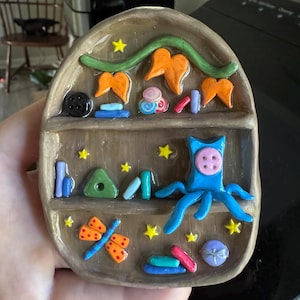 K&ouml;nnte beinhalten: Handgefertigtes Keramikregal mit verschiedenen bunten Miniaturdekorationen. Das Regal zeigt einen blauen Oktopus, einen orangefarbenen Schmetterling und andere Formen. Gelbe Sterne und ein schwarzer Knopf erg&auml;nzen das verspielte Design. Das Regal ist braun mit gl&auml;nzendem Finish.
