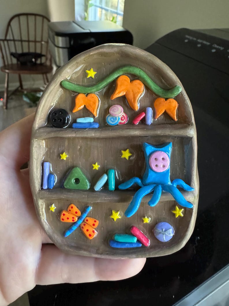 K&ouml;nnte beinhalten: Handgefertigtes Keramikregal mit verschiedenen bunten Miniaturdekorationen. Das Regal zeigt einen blauen Oktopus, einen orangefarbenen Schmetterling und andere Formen. Gelbe Sterne und ein schwarzer Knopf erg&auml;nzen das verspielte Design. Das Regal ist braun mit gl&auml;nzendem Finish.