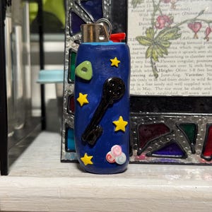 May include: A blue lighter with a key and star design. The lighter has a red button and a silver metal top. The design includes yellow stars, a green shape, and pink and white clouds.