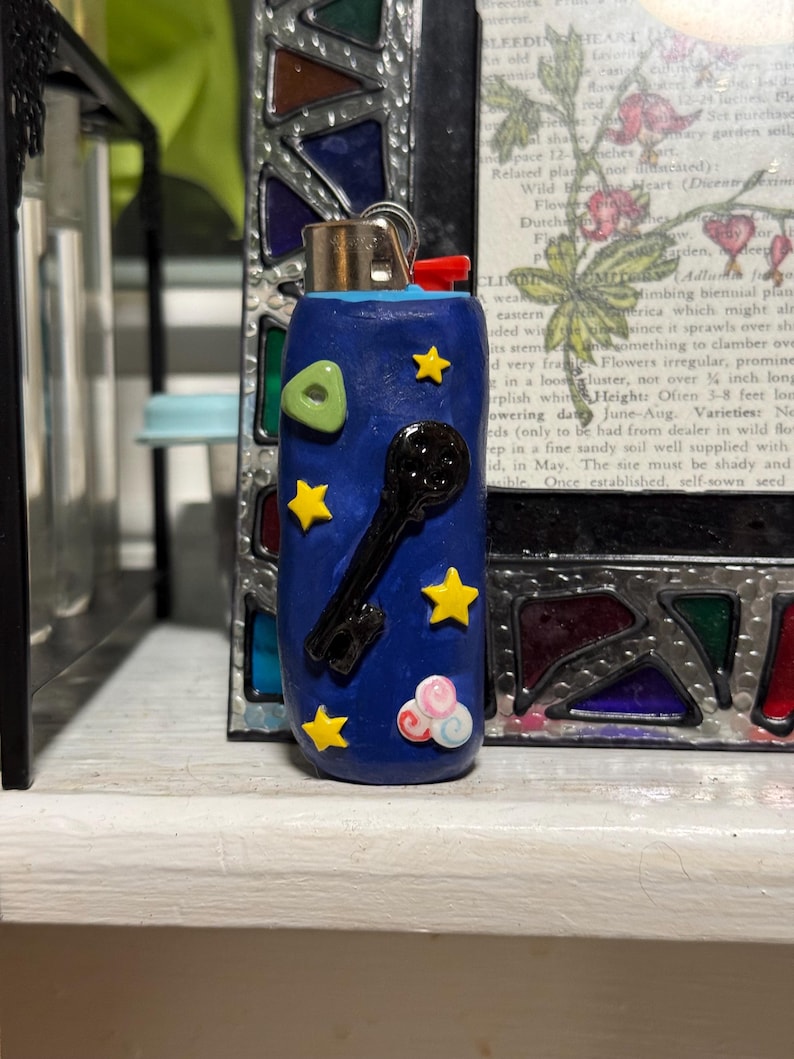 May include: A blue lighter with a key and star design. The lighter has a red button and a silver metal top. The design includes yellow stars, a green shape, and pink and white clouds.