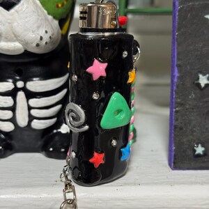 May include: Black lighter decorated with colorful stars, a green triangle, and silver accents. The lighter has a silver chain with a star-shaped charm. The lighter is on a white surface.