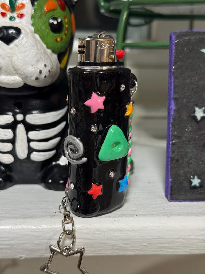 May include: Black lighter decorated with colorful stars, a green triangle, and silver accents. The lighter has a silver chain with a star-shaped charm. The lighter is on a white surface.