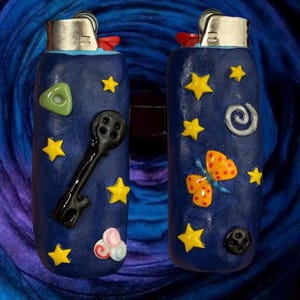 May include: Two navy blue lighters with decorative embellishments. One lighter features a black key and a green triangle, the other has an orange butterfly and a white spiral. Both have yellow stars.