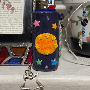 May include: A navy blue lighter with a decorative design. The lighter features a yellow circle with the words "Welcome Home" and colorful star accents. A silver star charm hangs from the side.
