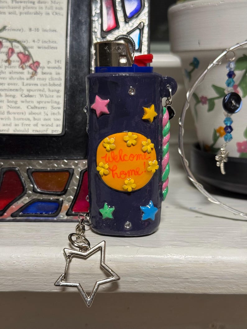 May include: A navy blue lighter with a decorative design. The lighter features a yellow circle with the words "Welcome Home" and colorful star accents. A silver star charm hangs from the side.
