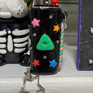 May include: Black lighter decorated with colorful star shapes, a green triangle, and small rhinestones. A silver star charm hangs from the side. The lighter has a red button and a silver metal top.