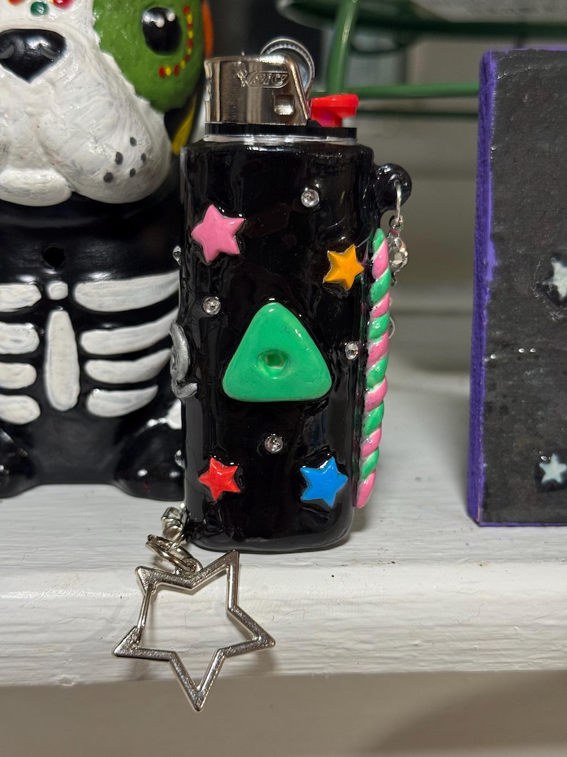 May include: Black lighter decorated with colorful star shapes, a green triangle, and small rhinestones. A silver star charm hangs from the side. The lighter has a red button and a silver metal top.