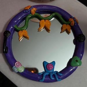 May include: A round, purple-framed mirror with whimsical clay embellishments. The frame is decorated with orange flower-like shapes, green vines, and a blue octopus-like creature. Additional details include a key, buttons, and a green triangle.