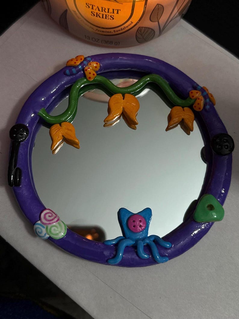 May include: A round, purple-framed mirror with whimsical clay embellishments. The frame is decorated with orange flower-like shapes, green vines, and a blue octopus-like creature. Additional details include a key, buttons, and a green triangle.