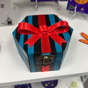 Pode incluir: Uma caixa hexagonal de madeira com listras verticais azuis e pretas, encimada por um laço de cetim vermelho. A caixa tem um fecho de metal e está em uma prateleira branca. Outros itens decorativos estão no fundo.