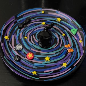 May include: A handmade trinket dish with a galaxy design. It has a swirling pattern in blue, purple, and black, with yellow star details. Small, colourful charms, including a red star, are scattered across the surface. A black, curved object is in the centre.