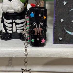 May include: Black lighter decorated with colorful stars and a bee design. A silver chain with a dragonfly charm and a star pendant hangs from the lighter. The lighter is on a white shelf next to a Day of the Dead dog figurine.