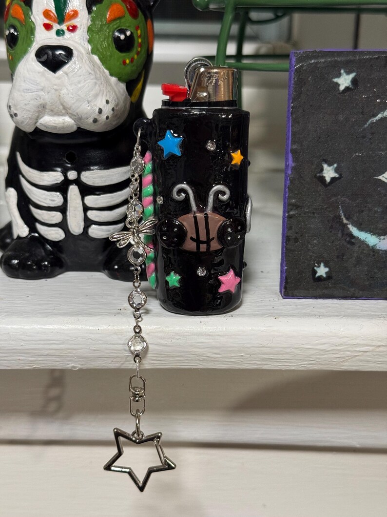 May include: Black lighter decorated with colorful stars and a bee design. A silver chain with a dragonfly charm and a star pendant hangs from the lighter. The lighter is on a white shelf next to a Day of the Dead dog figurine.