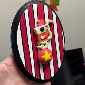 Puede incluir: Decoraci&oacute;n de pared ovalada con tem&aacute;tica de circo. El fondo presenta rayas rojas y blancas, con un borde negro. Un rat&oacute;n de dibujos animados con un atuendo rojo se equilibra sobre una bola roja con una estrella amarilla.