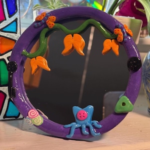 May include: A round, purple mirror with a decorative frame. The frame features orange flower-like shapes, green vines, and a blue octopus-like creature. Additional embellishments include a green triangle and a pink and green swirl.