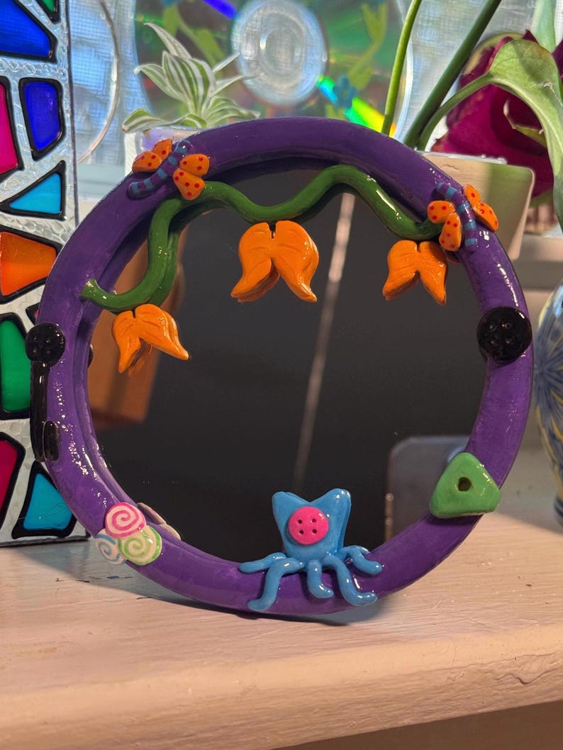May include: A round, purple mirror with a decorative frame. The frame features orange flower-like shapes, green vines, and a blue octopus-like creature. Additional embellishments include a green triangle and a pink and green swirl.