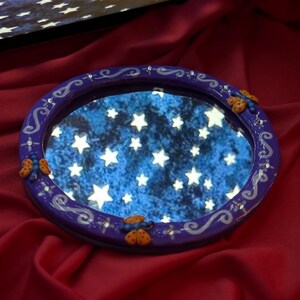 May include: A round, purple-framed mirror with a starry night sky reflection. The frame is decorated with silver swirls, small crystals, and orange butterfly accents. The mirror sits on a red satin surface.