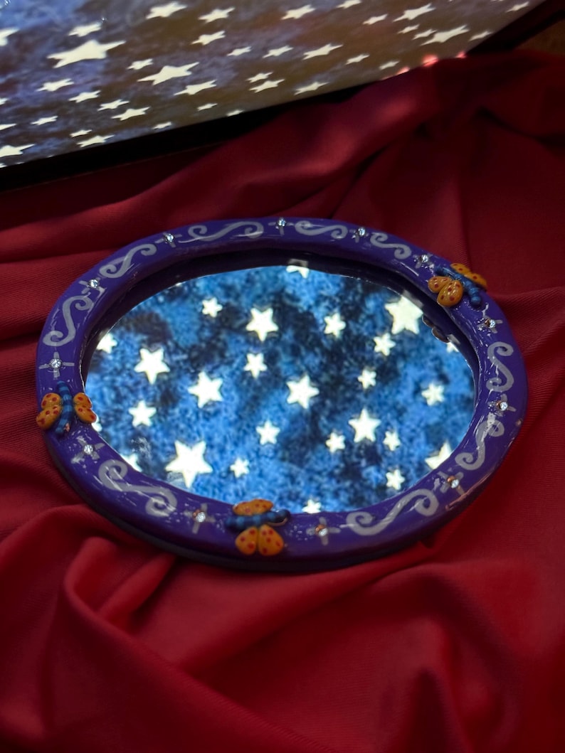 May include: A round, purple-framed mirror with a starry night sky reflection. The frame is decorated with silver swirls, small crystals, and orange butterfly accents. The mirror sits on a red satin surface.