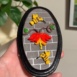 May include: An oval-shaped wall hanging featuring a brick-like background and a black frame. The artwork depicts a red and yellow flower with two yellow and black hummingbird figures. The piece is approximately 6 inches in height.