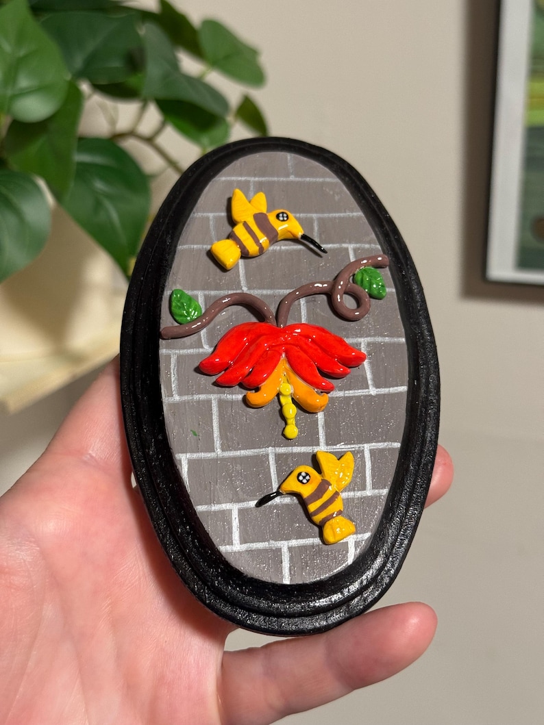 May include: An oval-shaped wall hanging featuring a brick-like background and a black frame. The artwork depicts a red and yellow flower with two yellow and black hummingbird figures. The piece is approximately 6 inches in height.