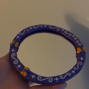 May include: A round, purple mirror with a decorative border. The border features white swirls, crosses, and small, colorful butterfly accents. The mirror is held in a hand.